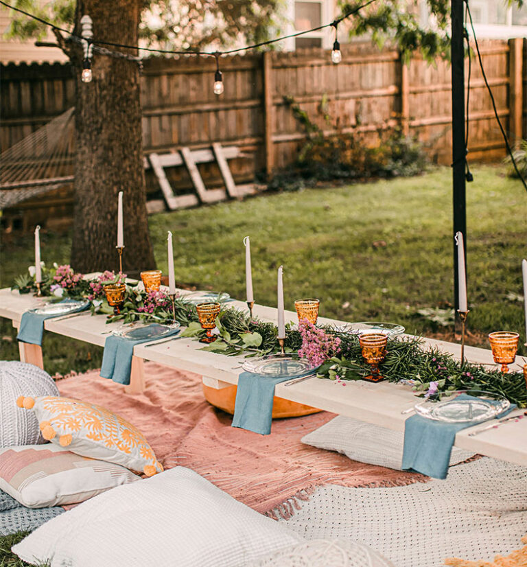 Low, lantern-lit outdoor dining setup on a long low table with candles, greenery, and colorful cushions on a yard blanket setup.