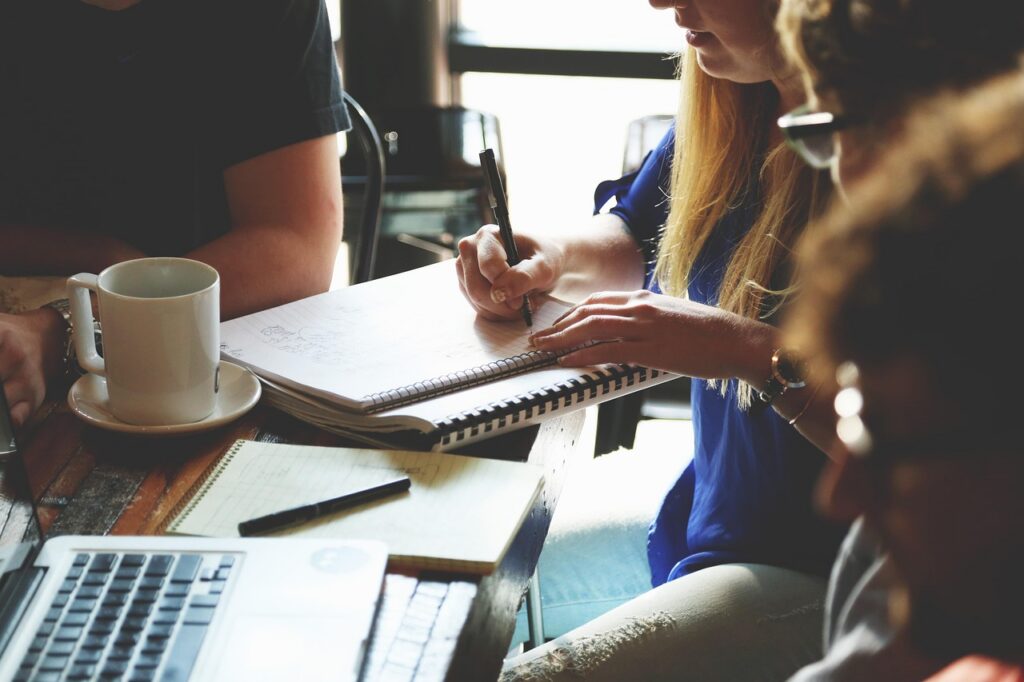 People collaborating at a wooden table; a woman writes in a spiral notebook beside a laptop and a coffee mug.