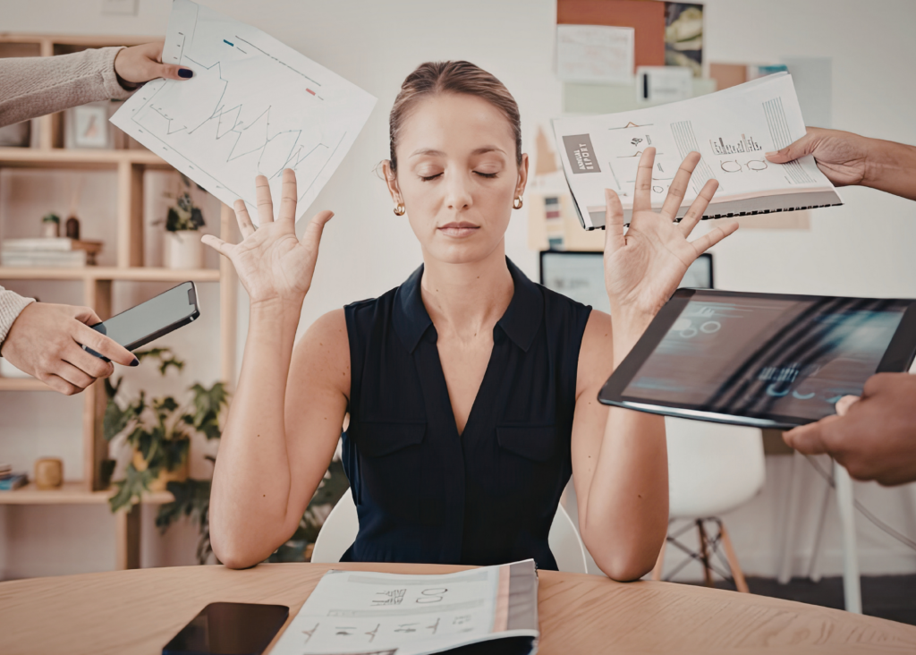 A woman sits with eyes closed while several colleagues hold up graphs and documents around her at a desk.