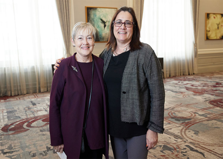 Two women stand side by side, smiling at the camera in a bright, formal room with patterned carpet and art on the wall.