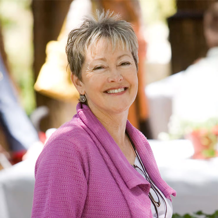 Smiling woman with short gray hair wearing a pink cardigan outdoors.
