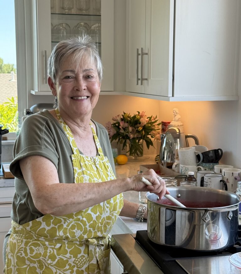 Older woman wearing a yellow floral apron stirs a red sauce in a stainless steel pot on the stove
